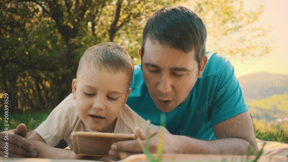 Father and son playing smartphone under sun light. Green field in park at sunny day mountains background Father and son playing smartphone under sun light. Green field in park at sunny day mountains background