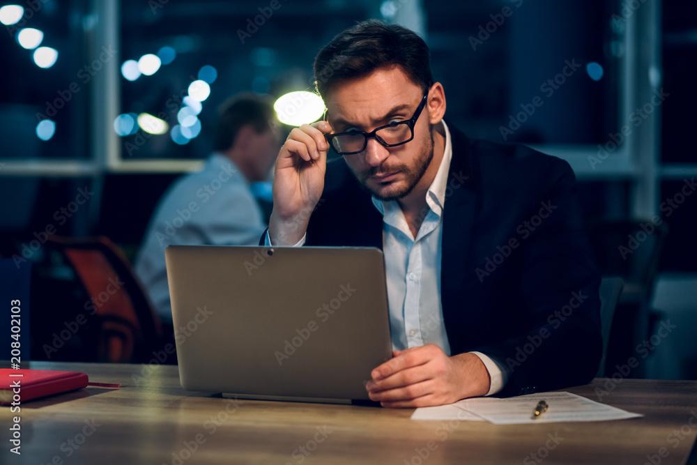 Bearded young businessman working at modern office. Business office worker wearing glasses sitting at wooden table using laptop for work.