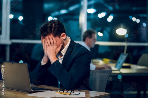 Tired man working late in office. Exhausted businessman sitting at office table with his face burried in his hands with laptop in front of him.