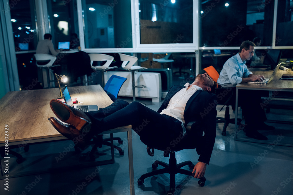 Guy lying in his chair asleep in office. Three businessmen working in office  late at night one of them sitting in his chair with his feet up on table  snoozing. Stock Photo