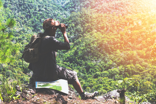 Young man with backpack and holding a binoculars,he is sitting on rock top of a mountain.