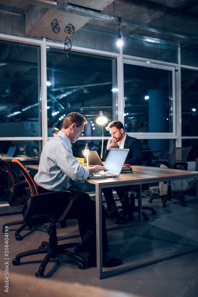 Couple of colleagues sitting at office table with laptops. Two serious businessmen working in middle of night in their modern office with glass windows. Couple of colleagues sitting at office table with laptops. Two serious businessmen working in middle of night in their modern office with glass windows.