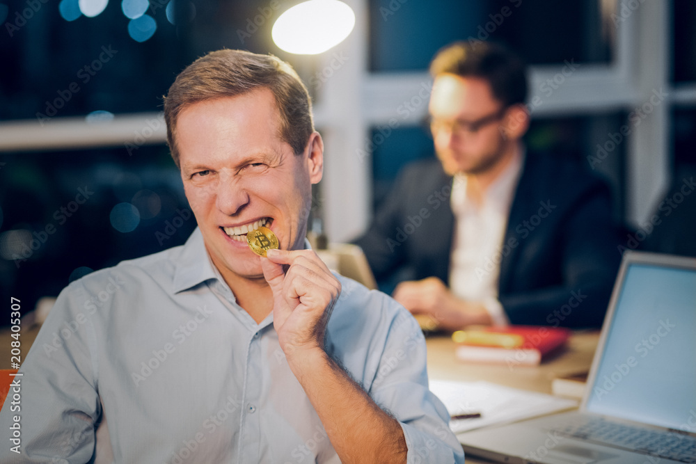 © Svyatoslav Lypynskyy - Funny mature man bites shiny golden bitcoin. Middle aged man wearing blue coloured shirt angrily biting golden bitcoin coin with his coworker working in background. © Svyatoslav Lypynskyy - Funny mature man bites shiny golden bitcoin. Middle aged man wearing blue coloured shirt angrily biting golden bitcoin coin with his coworker working in background.