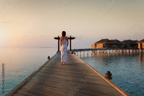 Elegante Frau im Abendkleid läuft bei Sonnenuntergang über einen Holzsteg auf den Malediven