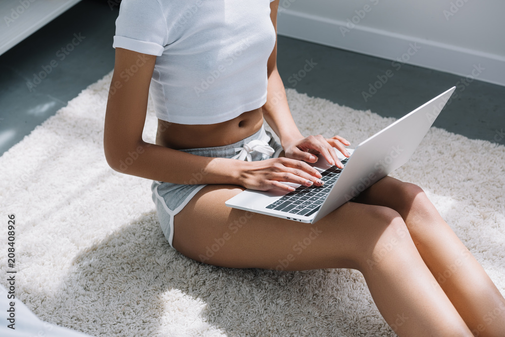 Naklejka premium cropped view of african american girl typing on laptop white sitting on floor