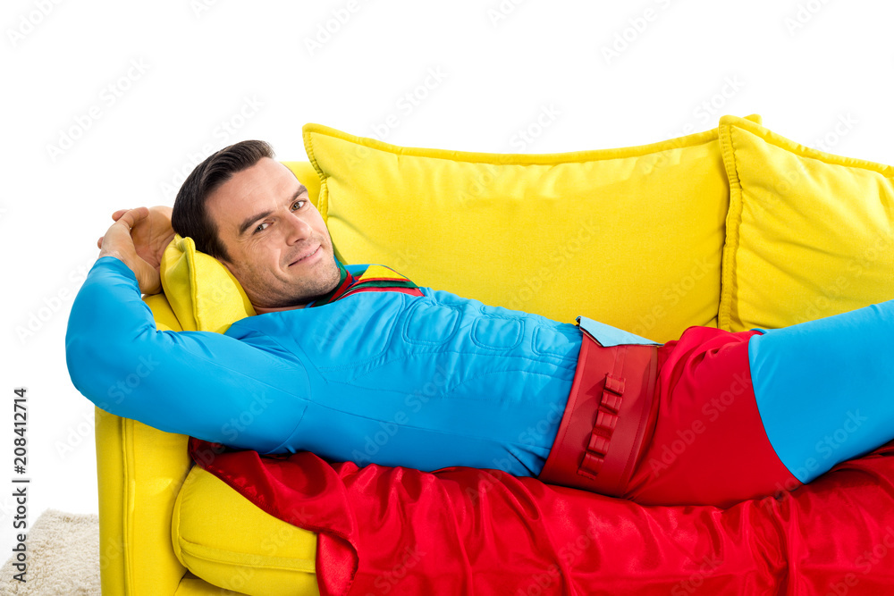 handsome man in superhero costume resting on couch and smiling at camera isolated on white