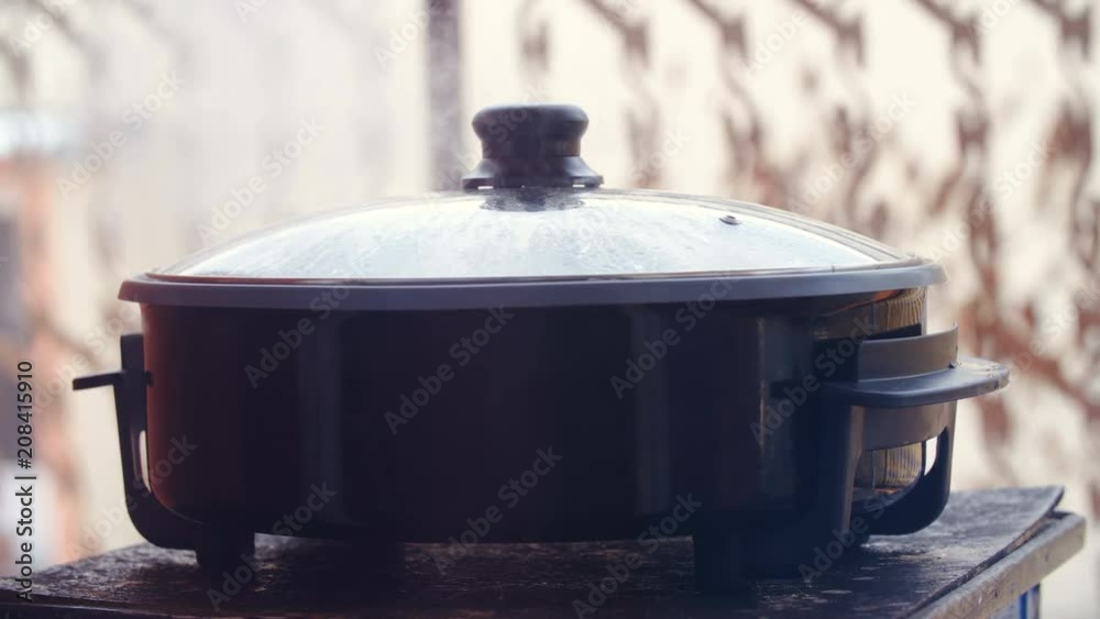 Making food in electrical cooking pot outdoors 4K. Long shot of a big cooking pot in focus while steam leaving from under the cover. Balcony fence in background.