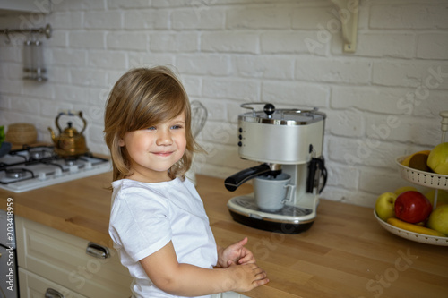 Horizontal shot of beautiful four year old baby girl in white casual t-shirt standing at kitchen counter, making coffee and smiling happily, feeling helpful and responsible. Children, kids and cooking