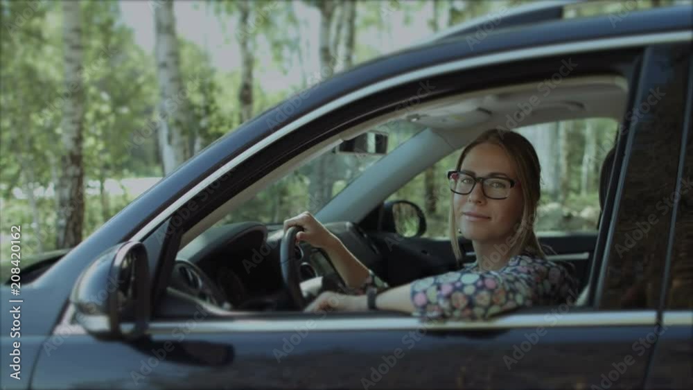 Portrait of smiling charming female driver in eyeglasses holding hand on steering wheel and looking out car window during summer vacation road trip. Pretty woman posing in luxury car during journey. Portrait of smiling charming female driver in eyeglasses holding hand on steering wheel and looking out car window during summer vacation road trip. Pretty woman posing in luxury car during journey.