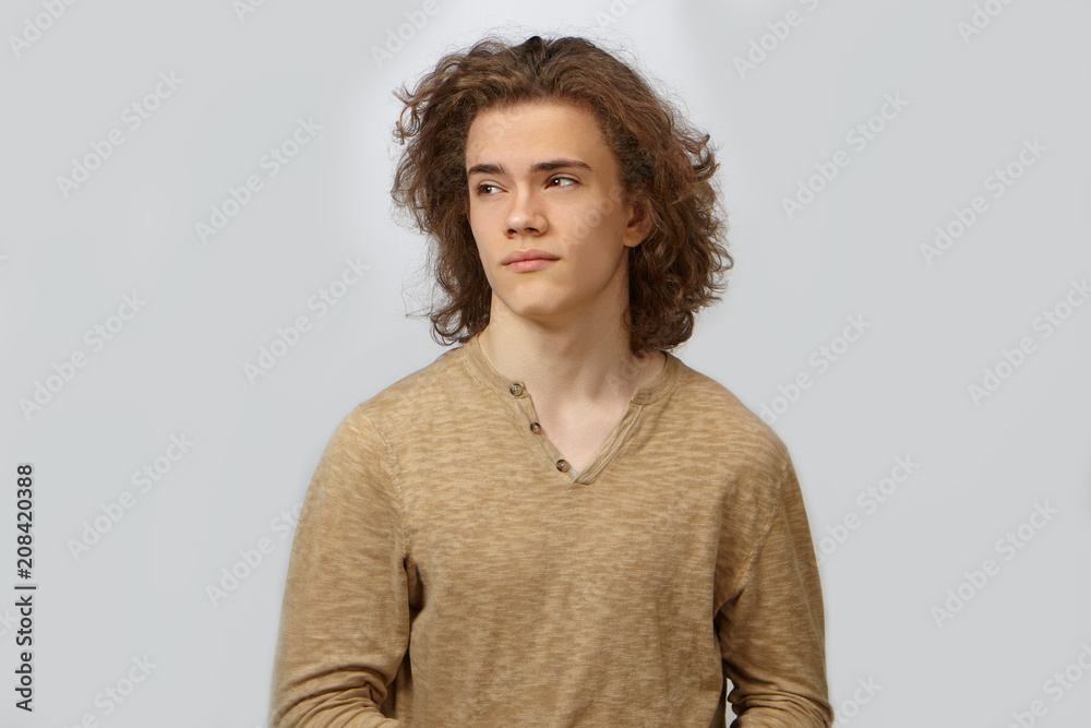 Portrait of attractive young European guy with smooth face, brown eyes and curly hairdo posing at blank studio wall, looking sideways, dressed in beige shirt. People, style and fashion concept Portrait of attractive young European guy with smooth face, brown eyes and curly hairdo posing at blank studio wall, looking sideways, dressed in beige shirt. People, style and fashion concept