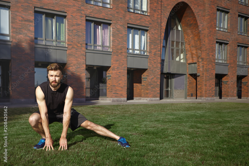 Sports, fitness, wellbeing, endurance and determination concept. Confident young Caucasian male jogger focused on training, doing side lunge on green grass, stretching legs before cardio exercise