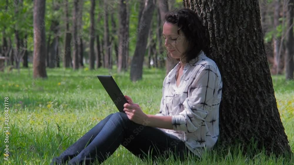 The girl is sitting under a tree with a tablet. A woman in a park with a gadget. The girl is sitting under a tree with a tablet. A woman in a park with a gadget.