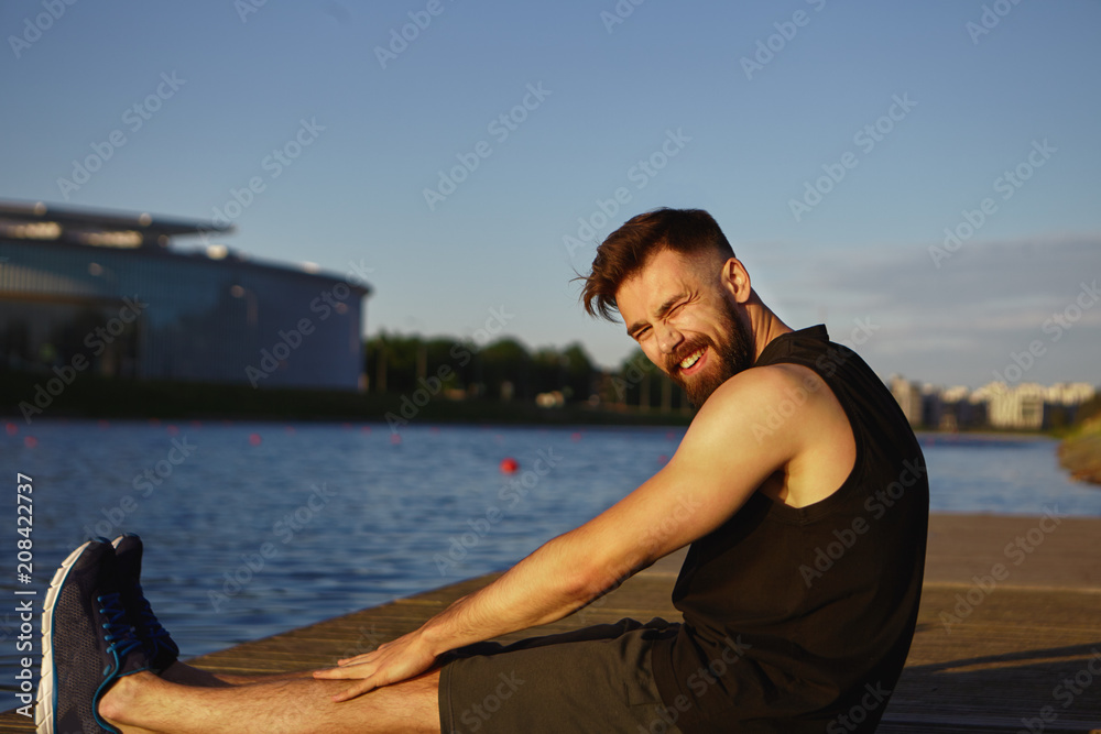 Outdoor shot of attractive bearded young Caucasian male athlete wearing stylish sports clothes sitting by river, squinting eyes because of bright sunshine, stretching legs after cardio morning routine