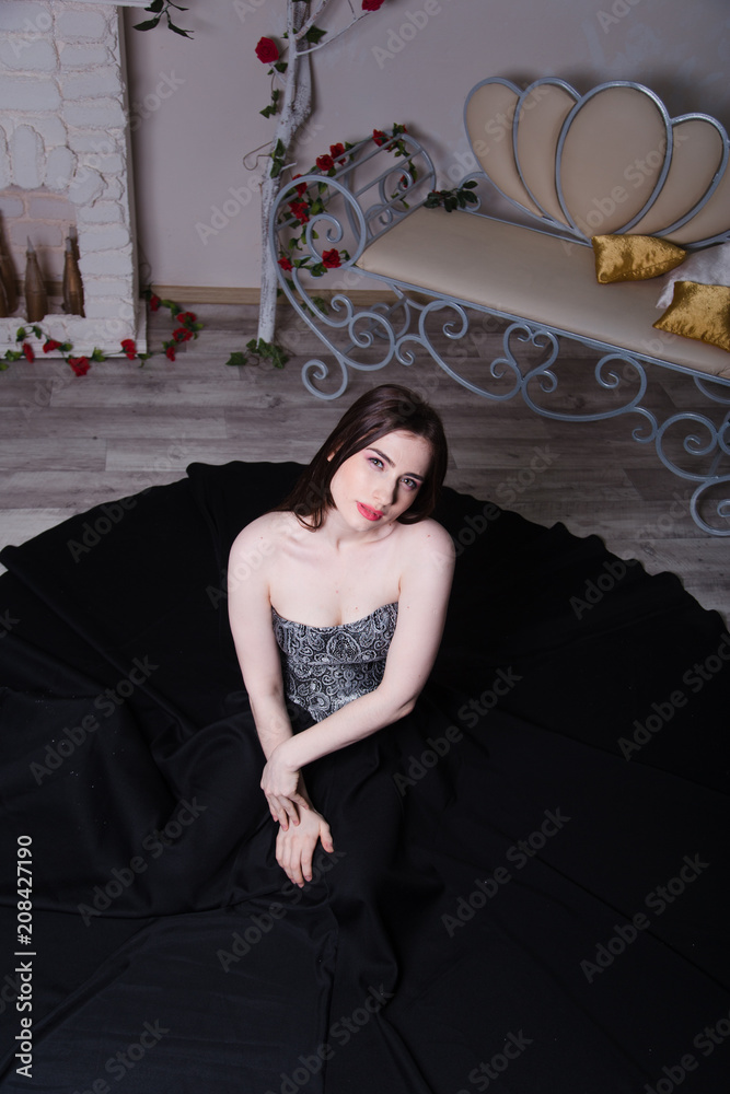 Fototapeta premium Beautiful girl in black long dress in white interior, model woman sits on the floor with white parquet.