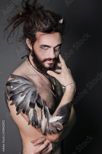 The image for halloween. A brutal guy with dreadlocks on a gray background. The guy with a thick beard. Hand the bird's wing. Feathers on hand. Fantasy