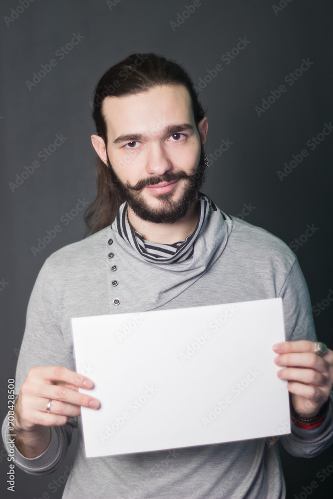 A bearded guy. Hipster model. fashion portrait in studio on a gray background with a piece of white paper in hands A bearded guy. Hipster model. fashion portrait in studio on a gray background with a piece of white paper in hands