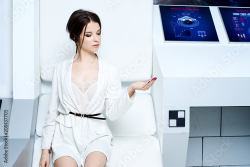Futuristic white interior reminiscent of a laboratory or a spaceship. Beautiful brunette in white clothes with blue eyes posing for the camera