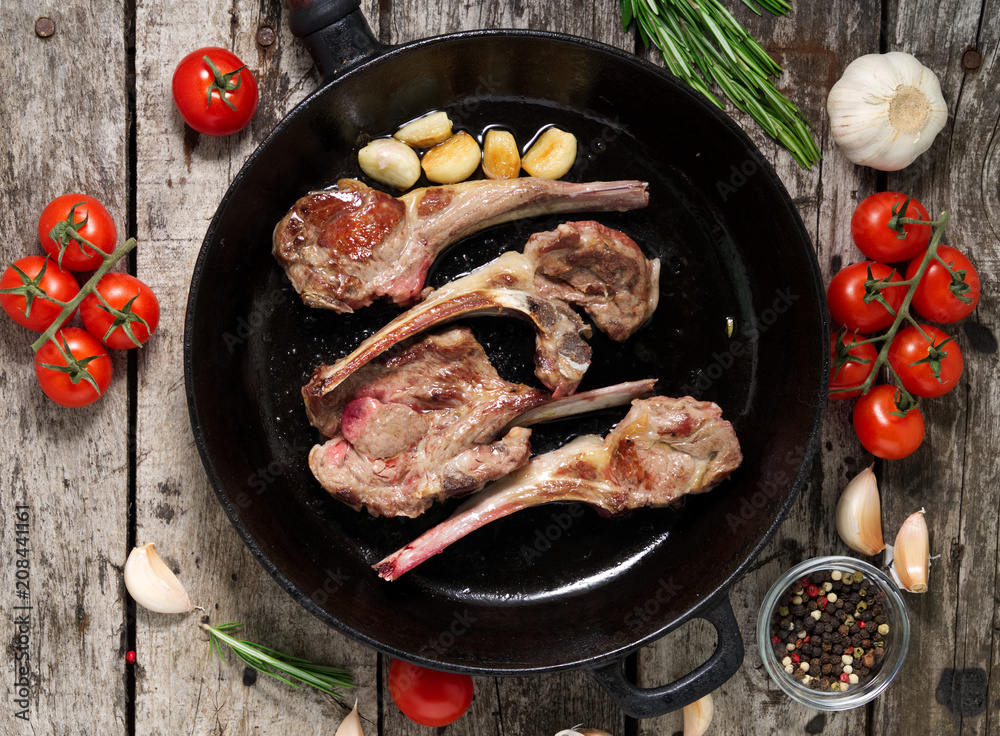 Roasted lamb ribs with rosemary, tomatoes and garlic on pan on dark old rustic wooden background, top view Roasted lamb ribs with rosemary, tomatoes and garlic on pan on dark old rustic wooden background, top view
