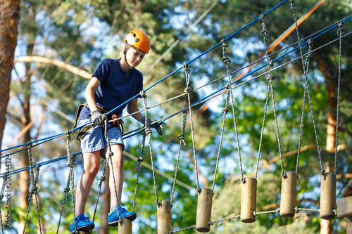 Active sporty kid in helmet doing activity in adventure park with all climbing equipment. Active children climb on the trees and having fun outdoors. Summer camp.