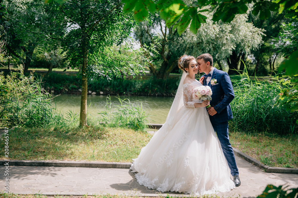 A stylish man in a suit in a cage hugs his beloved and buries his face in her blond hair. The bride in a gorgeous dress smiles next to her husband against the background of old green trees and a pond. A stylish man in a suit in a cage hugs his beloved and buries his face in her blond hair. The bride in a gorgeous dress smiles next to her husband against the background of old green trees and a pond.