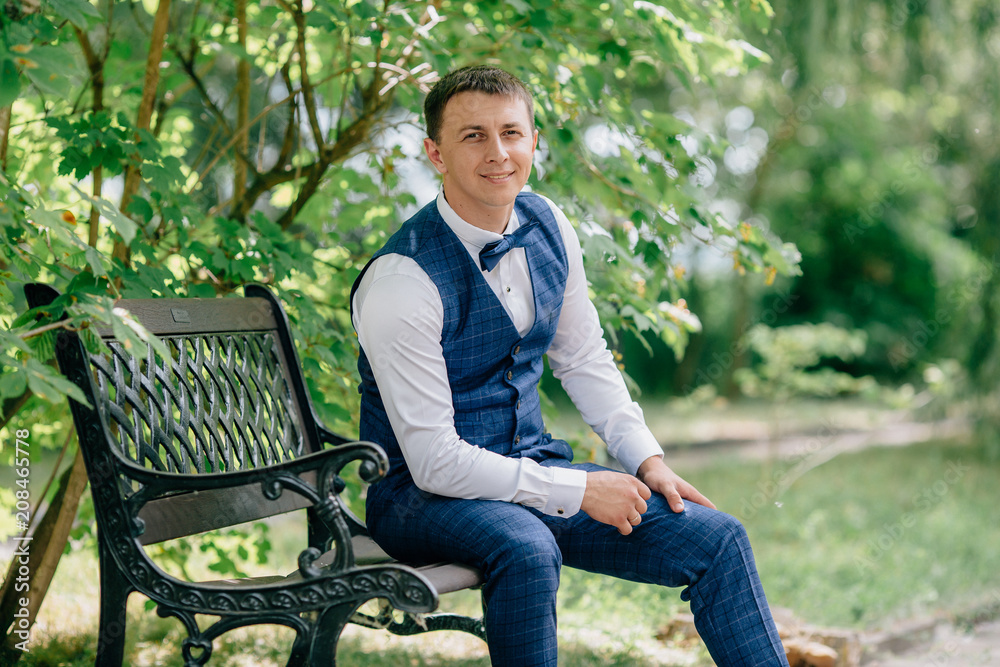 Portrait of business man in suit sitting in park on bench and smiling. The guy is getting ready for the wedding day, waiting for his bride. Portrait of business man in suit sitting in park on bench and smiling. The guy is getting ready for the wedding day, waiting for his bride.