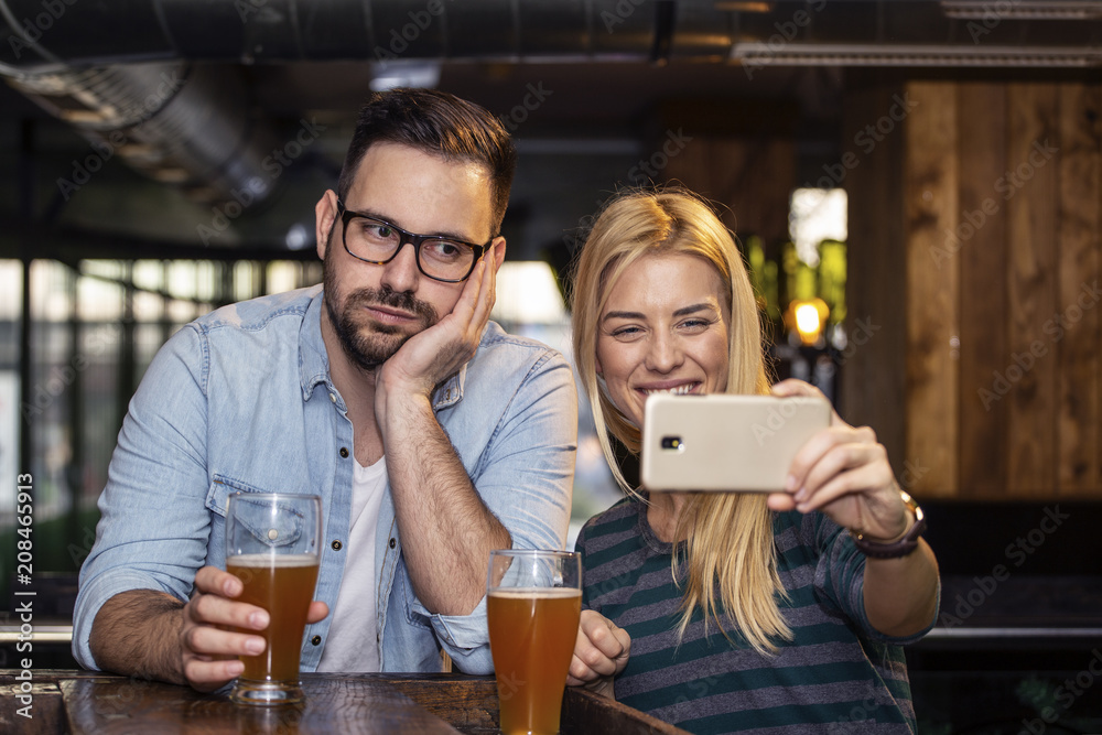 Fototapeta premium Young couple taking selfie by mobile phone on local pub and she smile while he is boring