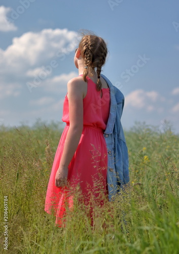Młoda kobieta, blondynka, uczesana w krótkie warkoczyki, stoi tyłem na łące, ubrana w letnią różową sukienkę bez rękawów, jeansowa kurtka zarzucona na ramię, relaksuje się, romantyczny widok