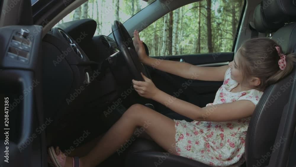 Joyful elementary age girl sitting in drivers seat and pretending to drive parents car. Carefree child having fun, playing in the car on driver seat during summer vacation road trip. Joyful elementary age girl sitting in drivers seat and pretending to drive parents car. Carefree child having fun, playing in the car on driver seat during summer vacation road trip.