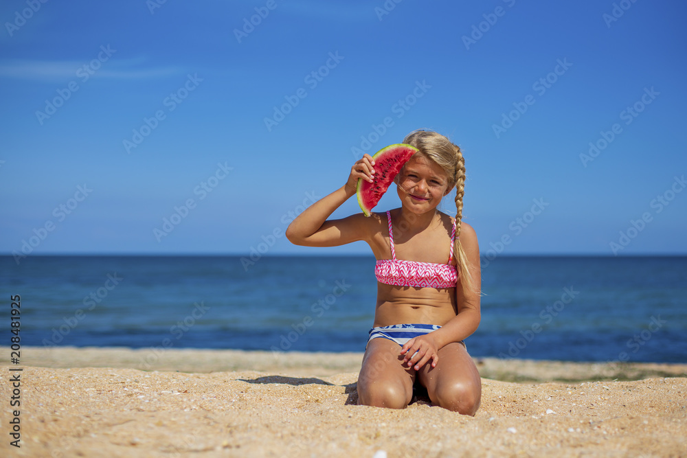 Portrait of a pretty blonde little girl in swimsuit eating watermelon on sandy beach near the sea on sunny day. Health eating. Sport and recreation	