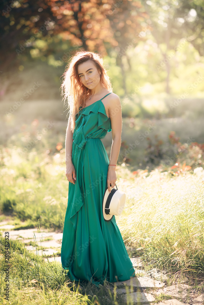 A young model girl in a long green dress and a beautiful hat walks along a path in a summer blooming garden in the background of a bright sun