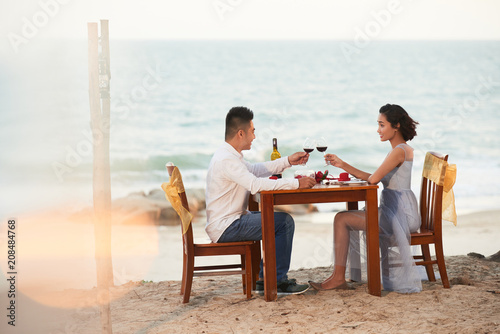 Profile view of joyful Asian couple toasting with red wine while celebrating Saint Valentines Day on beach of tropical island, picturesque seascape on background © DragonImages