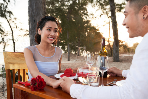 Cheerful Asian couple holding hands and looking at each other with toothy smiles while gathered together at beach restaurant and celebrating wedding anniversary