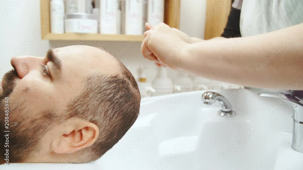 Person smear shampoo in hands and apply to man hair 4K. Close up slow motion of female person hands in focus applying shampoo to male head inside the sink at hairdresser salon. Person smear shampoo in hands and apply to man hair 4K. Close up slow motion of female person hands in focus applying shampoo to male head inside the sink at hairdresser salon.