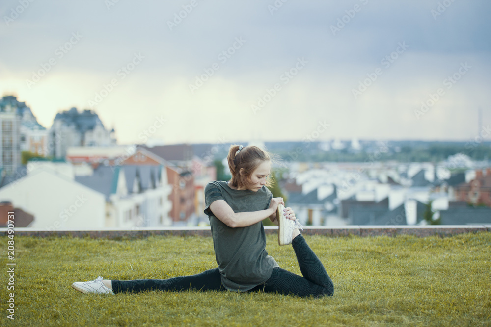 Young female acrobat on the grass performs twine, cityscape on background Young female acrobat on the grass performs twine, cityscape on background