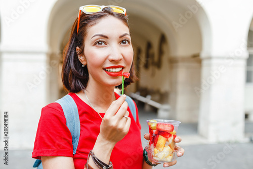 Attractive young woman eating fresh fruit salad while walking through the historic center of the European city