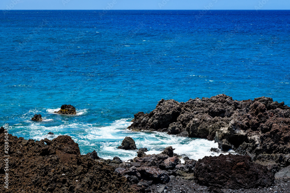 Bright blue Pacific Ocean with a rough black lava rock shoreline and crashing waves, Hawaii
