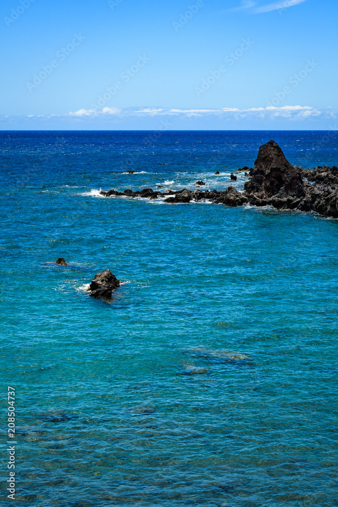 Obraz premium Bright blue Pacific Ocean with a rough black lava rock shoreline and crashing waves, and blue a sky with white clouds in the background, Hawaii 