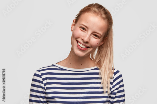 Happy female student with broad smile, has light hair combed in pony tail, recieves positive news, dressed in striped casual jacket, glad to meet with best friend, isolated on white background © wayhome.studio 
