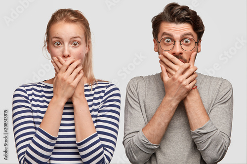 Shocked male and female teenagers cover mouth and look surprisingly at camera...