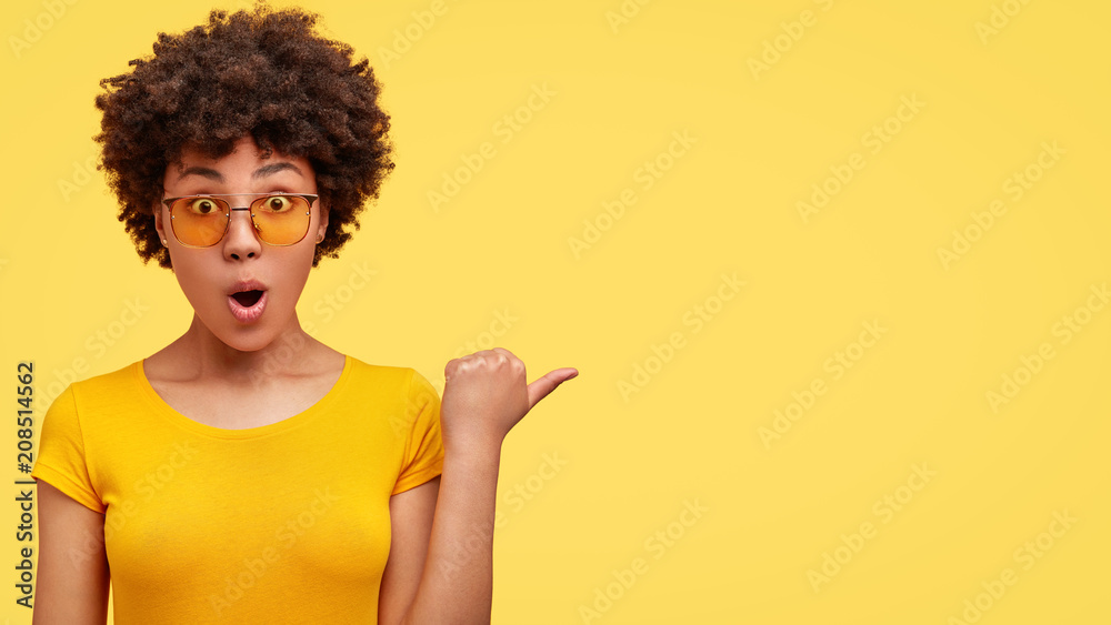 Horizontal shot of beautiful female with dark skin keeps mouth opened, wears casual t shirt and yellow shades, indicates at blank space with thumb, has stupefied expression. People, ethnicity concept - obrazy, fototapety, plakaty 