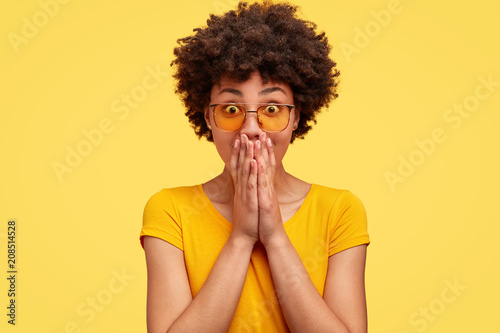 Portrait of amazed African American female covers mouth with surprisement  wears trendy shades  being stunned to recieve unexpected news  reacts on something. Momochrome and facial expressions