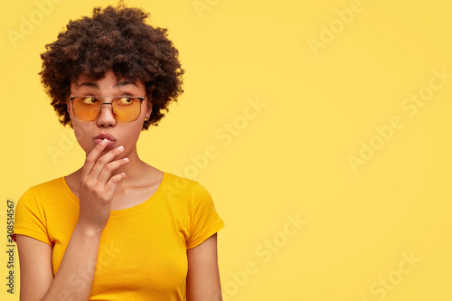 Pretty young dark skinned female contemplates about something and looks mysteriously aside, wears casual yellow t shirt and fashionable shades, isolated over blank wall. Monochrome, one tone