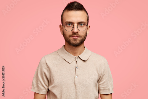 Serious handsome unshaven male student wonk in spectacles and brown t shirt, looks calm at camera, poses against pink background, ponders about graduation from university and future adult life © wayhome.studio 