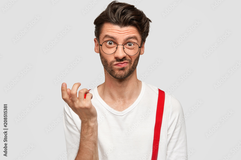 Fototapeta premium Frustrated puzzled male with European appearance, frowns face and raises hand, feels embarrassed after recieving difficult task at work, sees something not clear, isolated over white studio wall