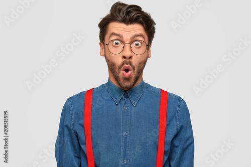 Photo of surprised hipster keeps mouth opened, feels puzzled and shocked, wears denim shirt with red suspenders, stares through spectacles, isolated over white background. It`s just unbelievablee! photo