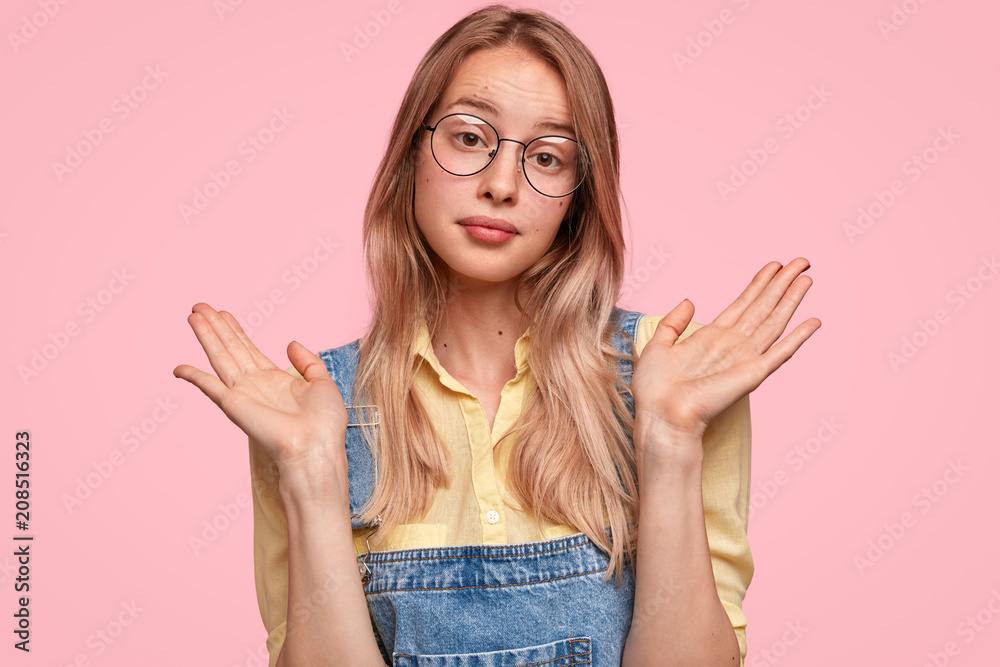 Clueless doubtful Caucasian female student in eyewear and jumpsuit, keeps palms raised, shruggs shoulders, being uncertain concerning something, isolated over pink background. Hesitation concept Clueless doubtful Caucasian female student in eyewear and jumpsuit, keeps palms raised, shruggs shoulders, being uncertain concerning something, isolated over pink background. Hesitation concept