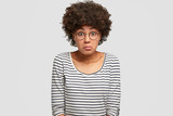 Studio shot of puzzled beautiful African American female has hesitant and frustrated expression, wears striped sweater, feels puzzlement, looks in bewilderment, sees something strange in front
