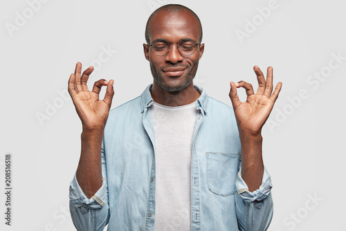 Portrait of pleased attractive African American guy makes okay gesture, has dark skin, smiles gently, eyes closed, wears denim shirt, isolated over white background. People, body language concept