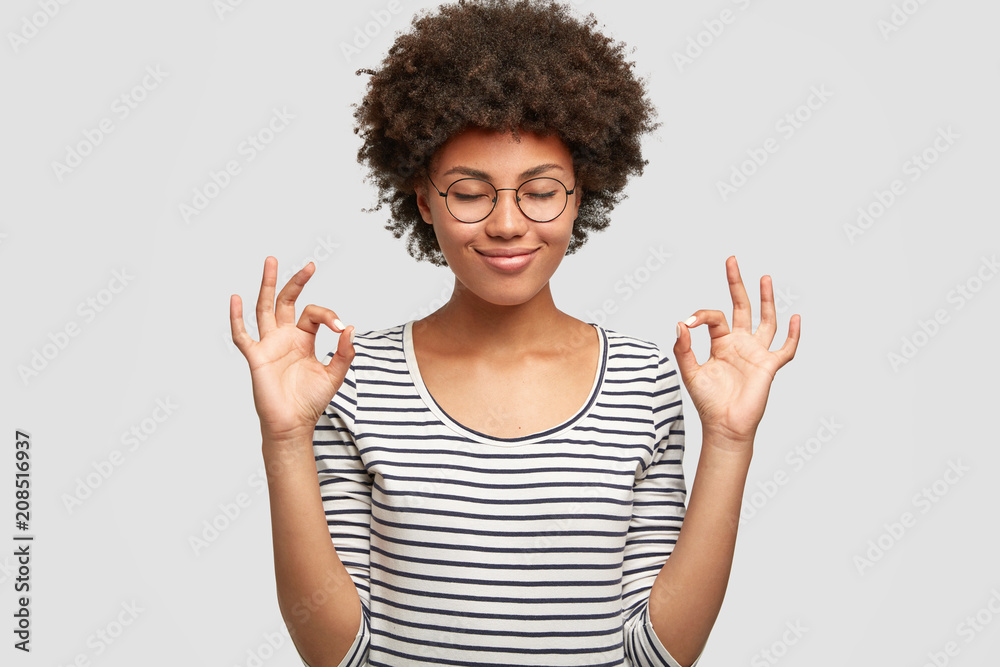 Horizontal shot of happy dark skinned female makes okay gesture, keeps eyes closed from pleasure, demostrates her agreement, has positve expression. Cheerful mixed race young woman poses indoor Horizontal shot of happy dark skinned female makes okay gesture, keeps eyes closed from pleasure, demostrates her agreement, has positve expression. Cheerful mixed race young woman poses indoor
