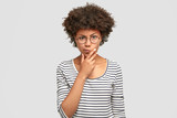 Horizontal shot of angry African American female curves lips and frowns eyebrows, keeps hand under chin, looks with discontent expression, feels frustrated, wears round glasses and striped sweater